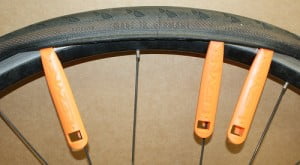 Start with two levers, near adjacent spokes. Then put a third lever next to them. Then continue, always putting the middle lever to one place further, until the tyre is off the rim (near half the circle).